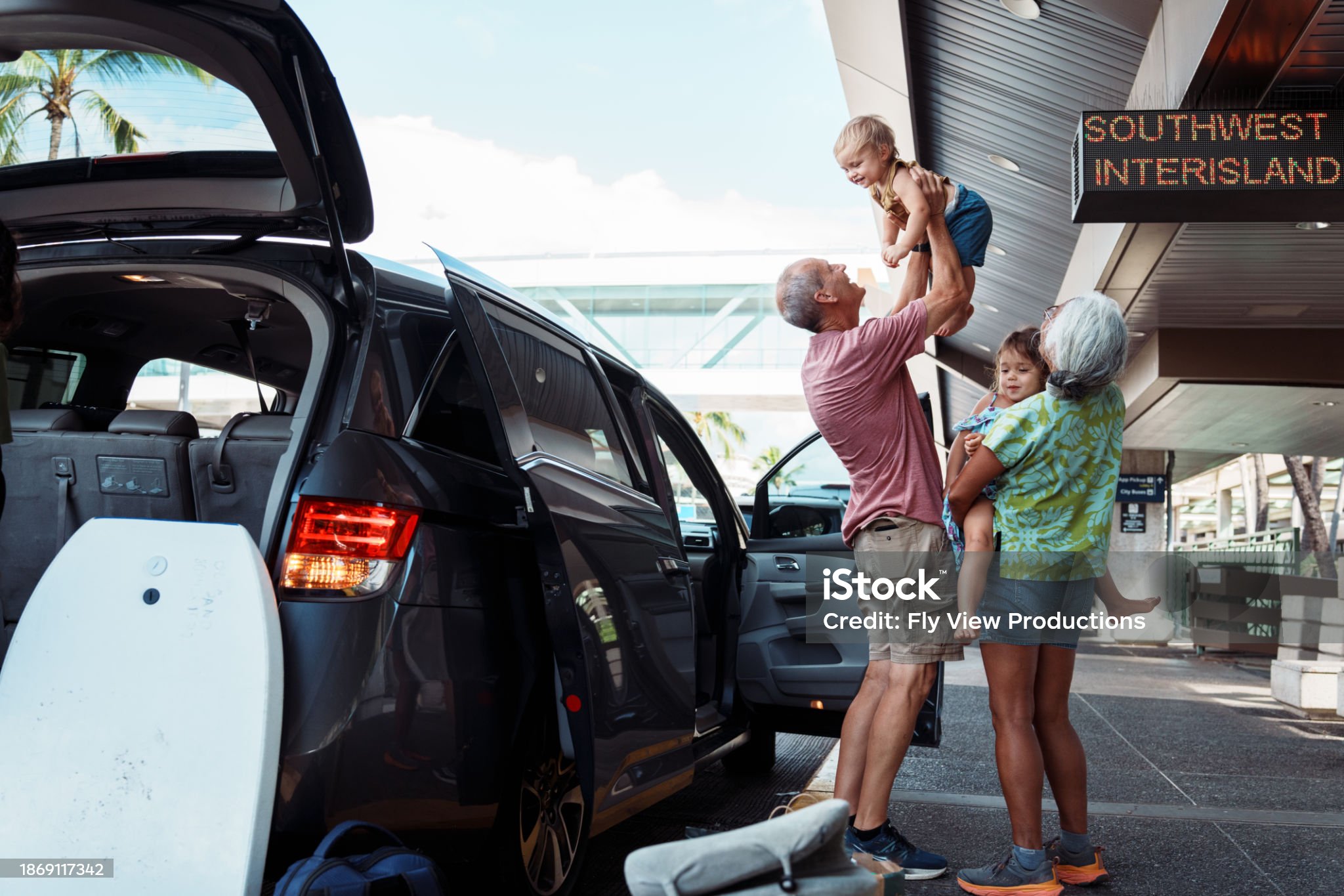 Grandparents lifting children at airport with luggage and black SUV