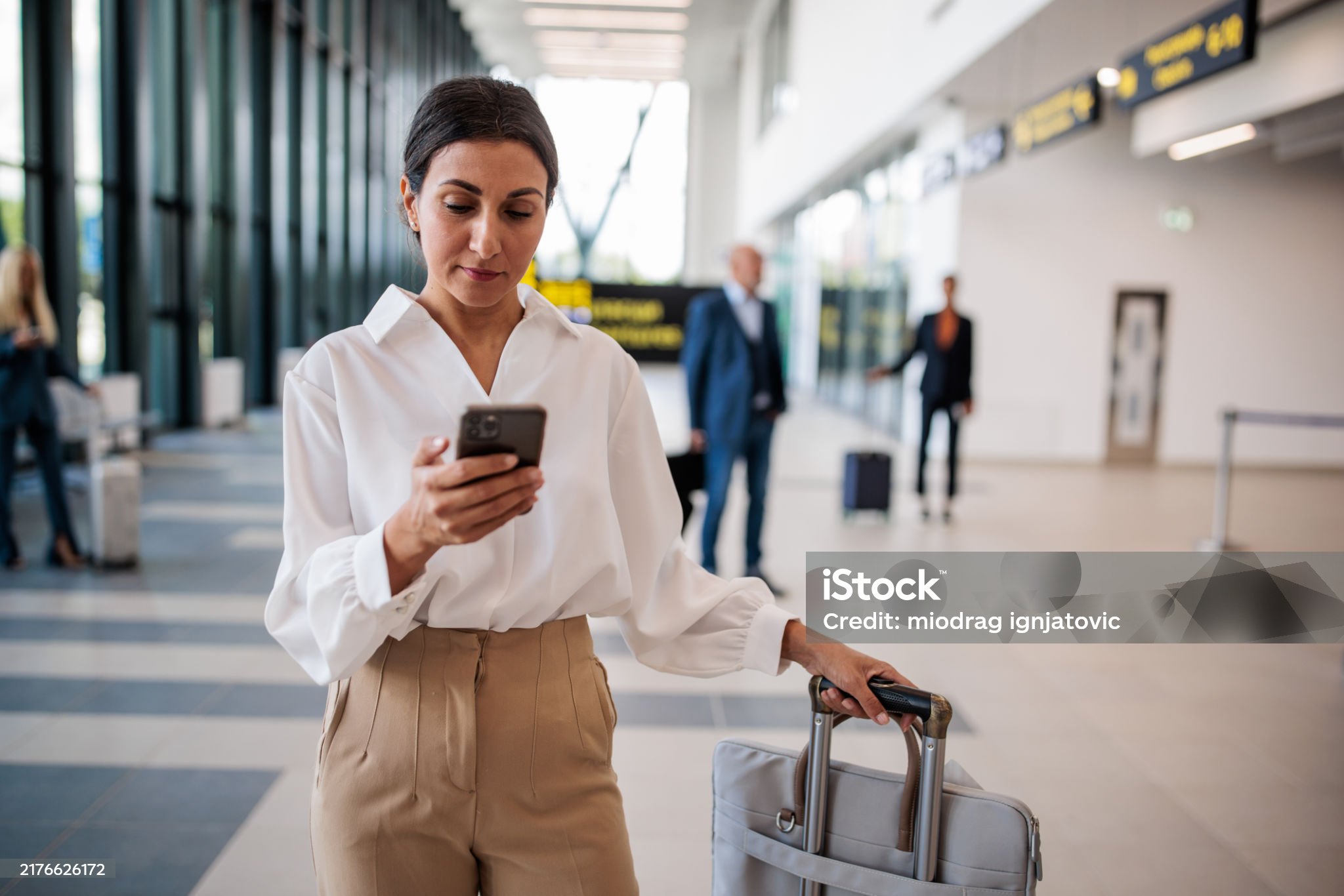 Business woman checking phone while walking through airport with luggage